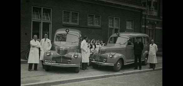Groepsfoto van het GG&GD-personeel in Nijmegen, naast twee nieuwe Chevrolet DP ambulances. Kees Rooijens is de derde van links.