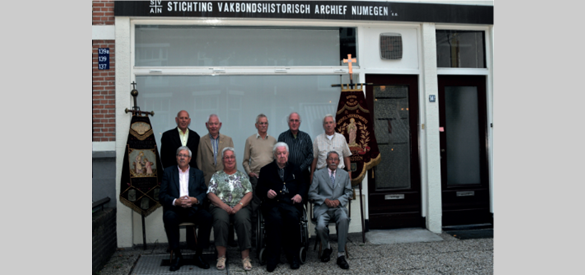 Het bestuur, geflankeerd door twee vaandels uit de collectie, zit voor de gevel van het stichtingsgebouw aan de Burghardt van den Berghstraat, 2008