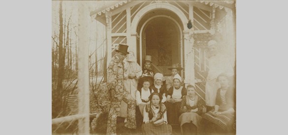 Groep gekostumeerde jongeren bij het prieel op het voorplein, met in bloemetjeskostuum en hoge hoed Goswin Eduard Lüps. Foto ca. 1900 – 1910. Bron: Gelders Archief, Arnhem