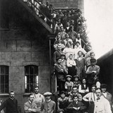 Personeel van schoenfabriek 'De Ooievaar' aan de Kerkstraat poseert op de trap van de fabriek in 1925. Onder de arbeiders zijn kinderen die net van de lagere school komen. Collectie G.G.Driessen.